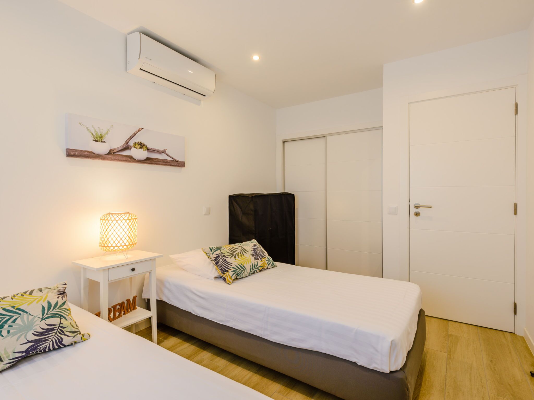 A single-bed room with a white bed, a bedside table with a lamp, a wall-mounted air conditioner, and a door.