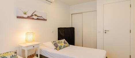 A single-bed room with a white bed, a bedside table with a lamp, a wall-mounted air conditioner, and a door.