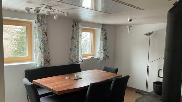 A dining area with a wooden table and black chairs, two windows with curtains, a ceiling fan, and a wood-burning stove.
