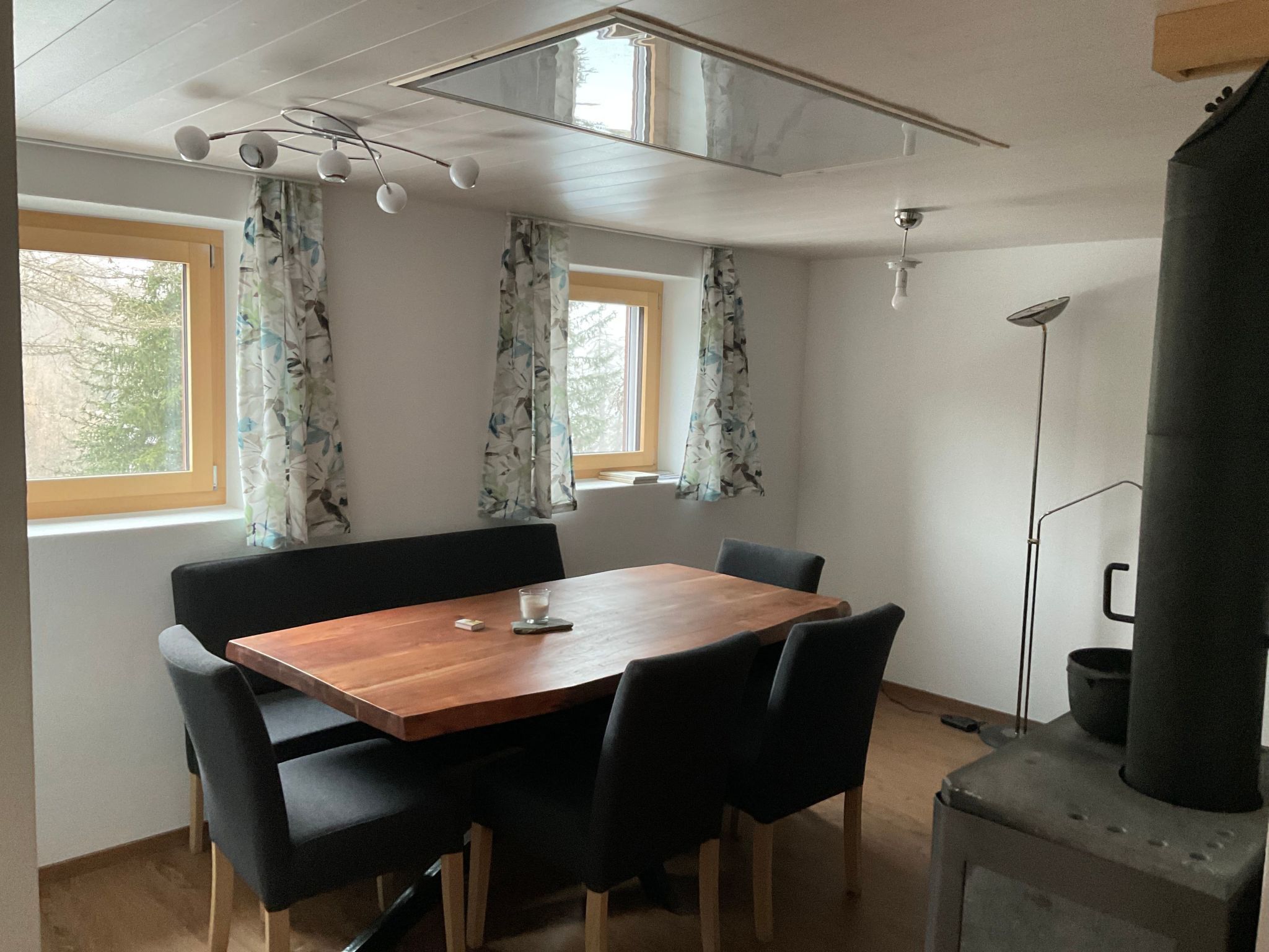 A room with a wooden table and chairs, two windows with curtains, a ceiling fan, and a wood stove.