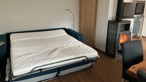 A bedroom with a bed, a wooden floor, a kitchenette with a stove, and a chair.