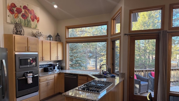 Una cocina moderna con armarios de madera, horno empotrado, microondas y estufa. Amplias ventanas ofrecen vistas a árboles.
