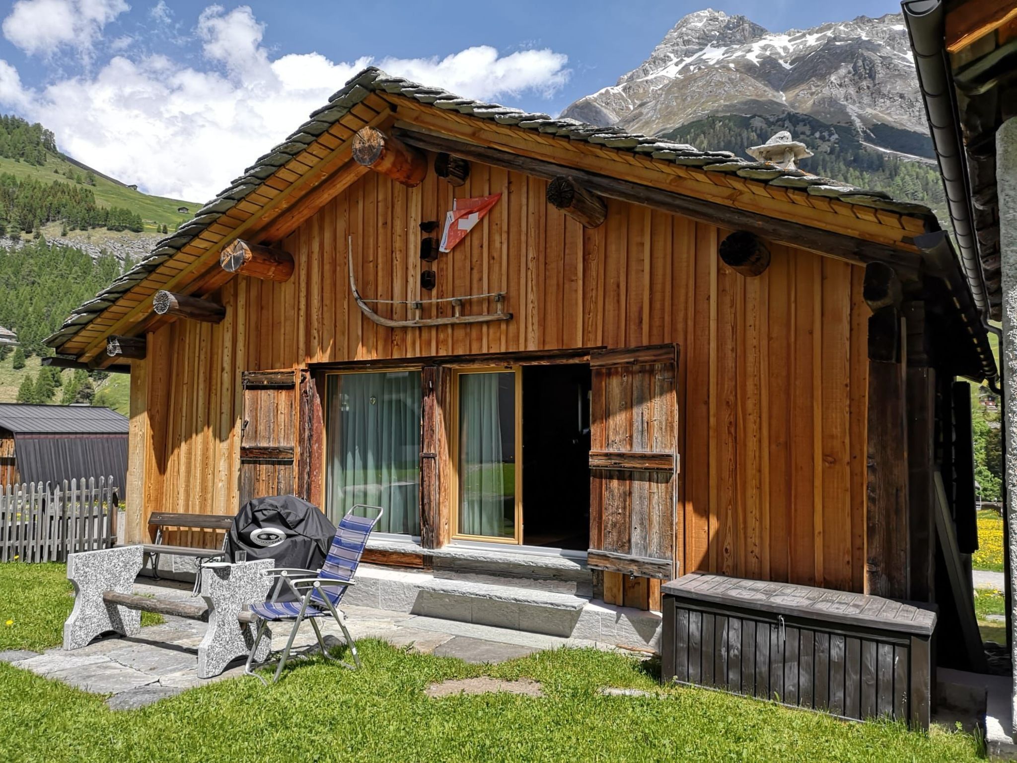 A wooden cabin with a stone bench and a grill outside, set against a mountainous backdrop.