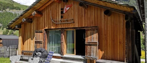 A wooden cabin with a stone bench and a grill outside, set against a mountainous backdrop.