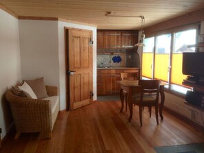 Ein gemütlicher Wohnbereich mit Holzboden, einem Rattansofa, einem Esstisch mit Stühlen und einer Küche mit Holdschränken und einem Fenster.