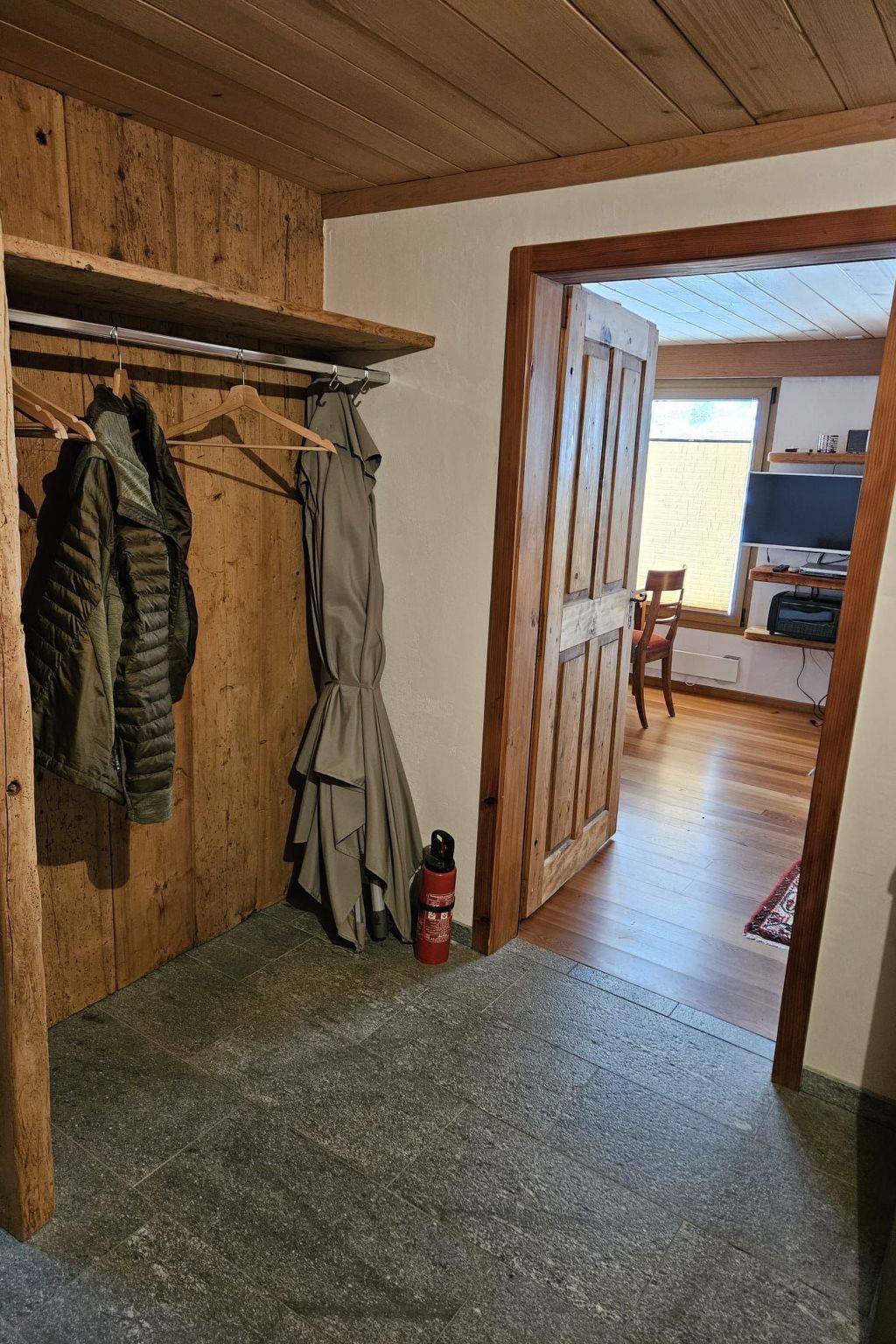 A room with a wooden ceiling, a coat rack with jackets, a door leading to another room, and a fire extinguisher on the floor.