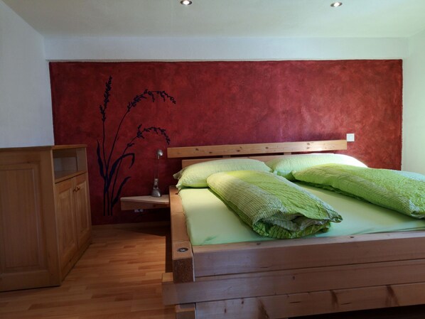 A bedroom with a wooden bed, green bedding, a wooden nightstand, and a red wall with a plant design.