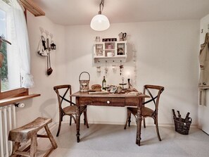 A cozy dining area with a wooden table, two chairs, a hanging light fixture, and a shelf with various items.