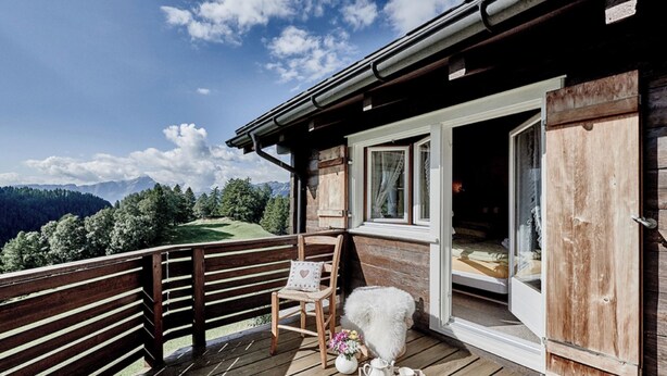 Una terraza de madera con una silla y una mesa pequeña, una cabaña de madera con puerta corredera y un paisaje montañoso al fondo.