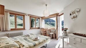 Un dormitorio con cama, escritorio, cómoda, radiador y ventanas grandes con vistas al exterior.