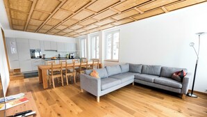 A modern living room with a wooden ceiling, a grey sectional sofa, a dining area with wooden chairs, and a kitchen in the background. - Ferienhaus Isla, (Nufenen), by Interhome (Nufenen)