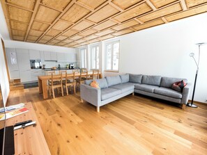 A modern living room with a wooden ceiling, a grey sectional sofa, a dining area with wooden chairs, and a kitchen in the background. - Ferienhaus Isla, (Nufenen), by Interhome (Nufenen)