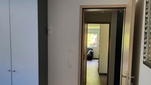 Un pasillo estrecho con una puerta que da a una habitación con una cama. Hay una pequeña mesa de madera e interruptor de luz en la pared.