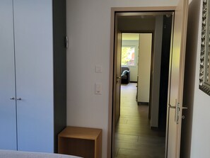 Ein schmaler Flur mit einer Tür zu einem Zimmer mit einem Bett. An der Wand befindet sich ein kleiner Holztisch und ein Lichtschalter.