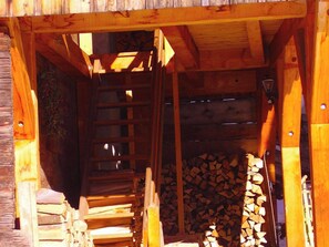 A wooden cabin interior with a staircase leading to a loft area, a stack of firewood, and a metal lantern. - Haus Nicca, (Splügen), by Interhome (Splügen)