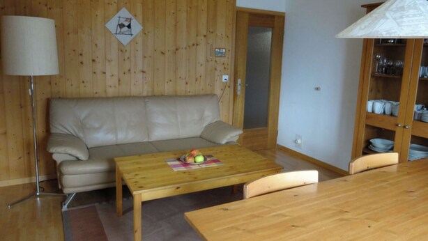A living room with a wooden wall, a beige sofa, a wooden coffee table with fruit, a lamp, and a cabinet with dishes.