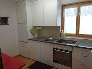 A kitchen with white cabinets, a double sink, a stove, and a window with lace curtains. - Tgea Beala Vesta, (Lohn). by Interhome (Lohn GR)