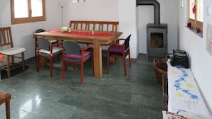 Un comedor con mesa y sillas de madera, suelo de baldosas, un calentador fijado a la pared y una ventana con persianas.