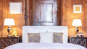 A bedroom with a wooden door, two bedside tables with lamps, a bed with white bedding, and decorative pillows.