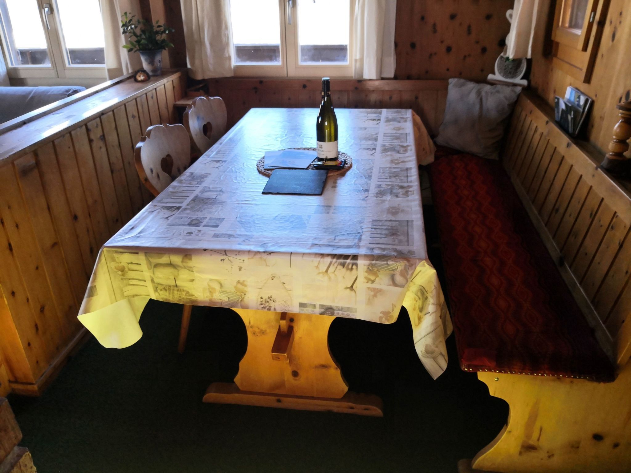 A wooden table with a newspaper-covered surface, a bottle of wine, and a bench with a cushion.
