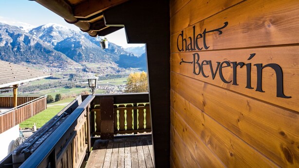 A wooden sign with 'Chalet Beverin' written on it, a wooden balcony, and a mountainous landscape in the background.