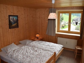 Een houten kamer met een bed, een nachtkastje, een lamp, een raam met uitzicht en een schilderij aan de muur.
