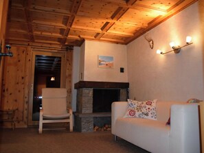 A cozy room with a stone fireplace, wooden ceiling beams, a white sofa, and a chair. - Wohnung Stücklihaus, (Sufers), by Interhome (Sufers)
