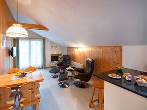 Una sala moderna con comedor, paredes de madera, chimenea y cocina al fondo.