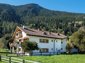 A two-story house with a balcony, surrounded by a wooden fence, set against a mountainous backdrop.