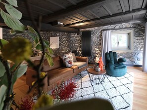 Un salon chaleureux avec un mur en pierre, un plafond en bois, un canapé marron, un fauteuil turquoise et une petite table agrémentée d’objets décoratifs.