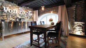 A rustic kitchen with a stone wall, wooden ceiling beams, a wooden dining table with chairs, and a stone fireplace.