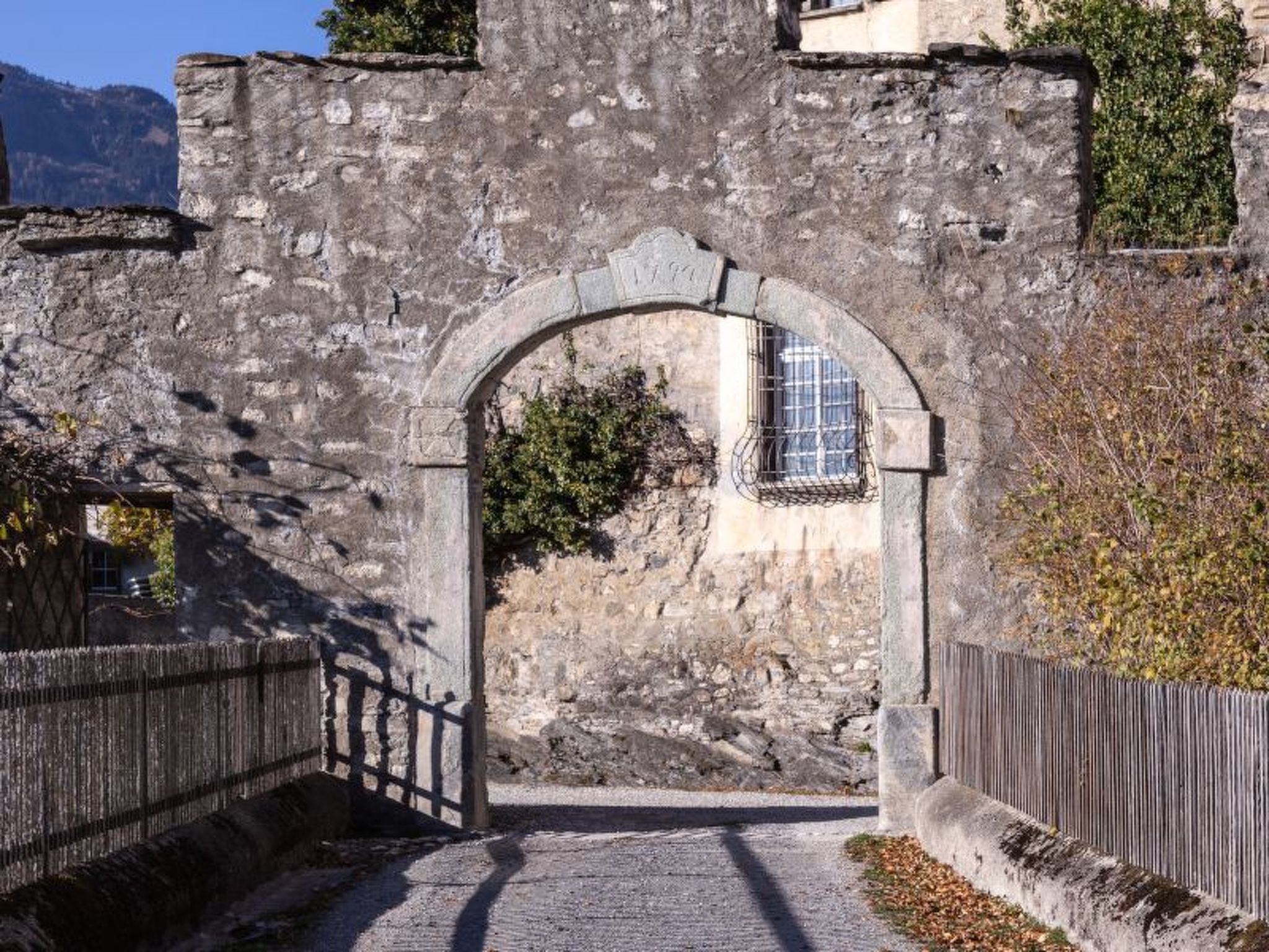 Un arco de piedra que conduce a un edificio con ventana, rodeado de un muro de piedra y una valla de madera.