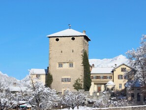 Un edificio histórico con torre y techo nevado, rodeado de edificios tradicionales con numerosas ventanas bajo un cielo azul despejado.