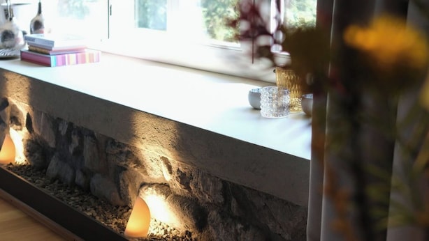 A room with a stone ledge, a window with a view of greenery, and a vase with flowers.