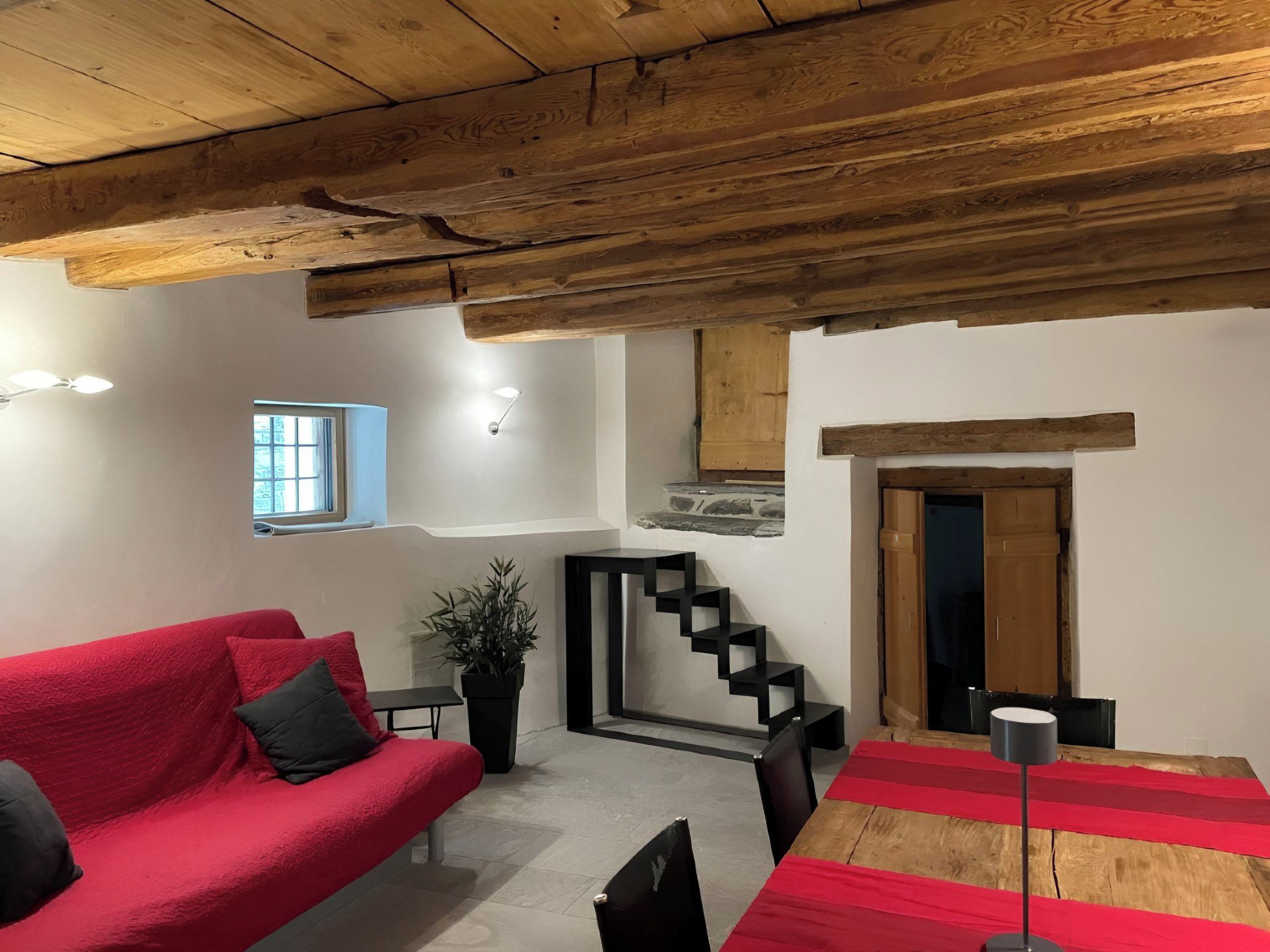 A cozy living space with a red sofa, a small table, a potted plant, a staircase, and a wooden ceiling.