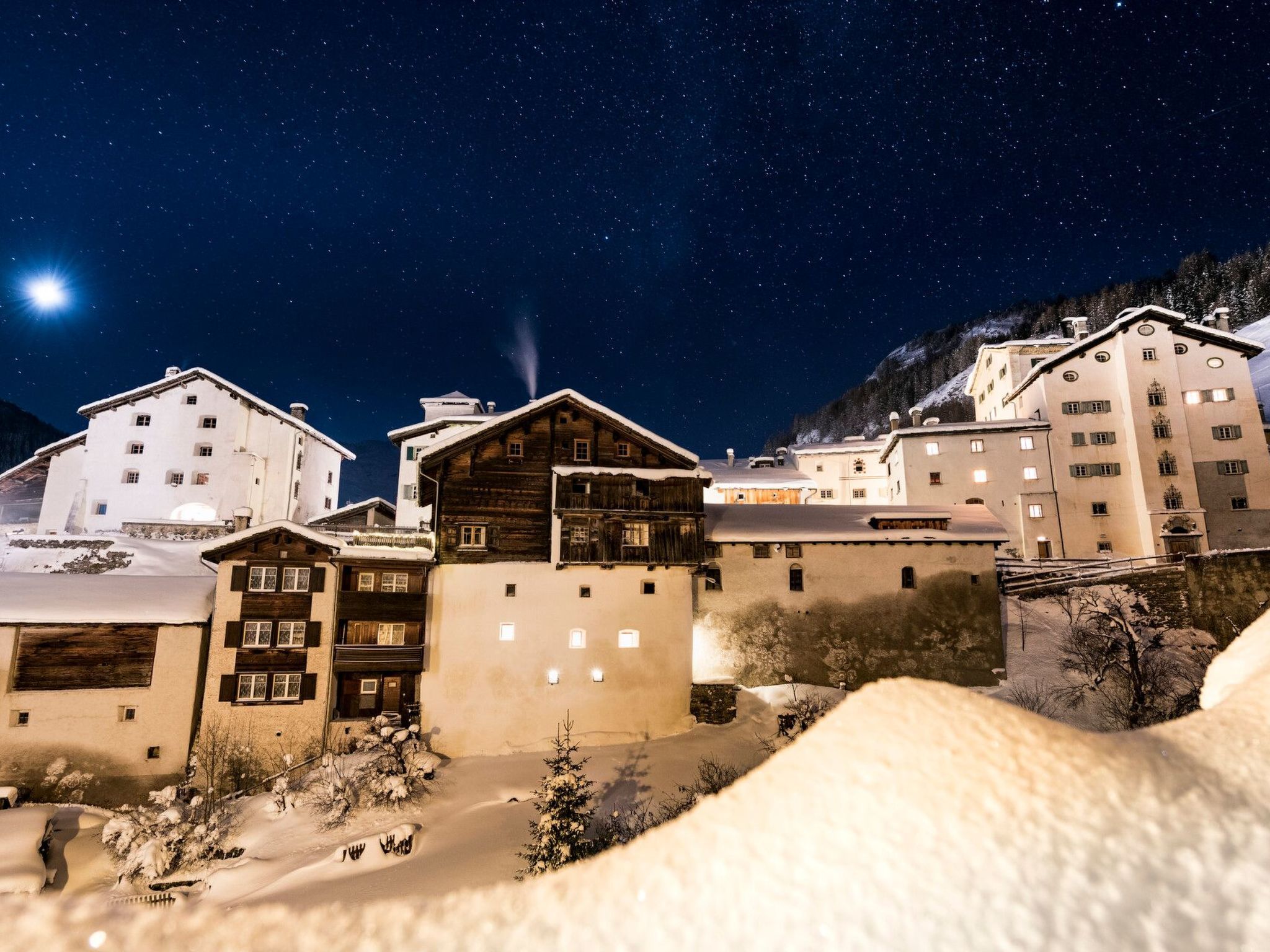 Een bergstad bedekt met sneeuw 's nachts, met verlichte gebouwen en een sterrenhemel.