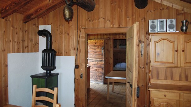 Interior de una cabaña de madera con una mesa de comedor, sillas, estufa, cama y un armario.