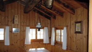 Interior de una cabaña de madera con una mesa de comedor, sillas, ventanas con cortinas y una lámpara colgante.