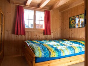 A wooden cabin bedroom with a bed, two windows with red curtains, and a painting on the wall. - Ferienhaus beim Kulturtenn, (Obermutten), by Interhome (Thusis)