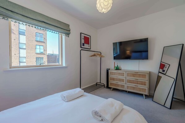 Une chambre d’hôtel équipée d’un lit, d’une télévision, d’une commode, d’un miroir et d’une fenêtre donnant sur un immeuble.