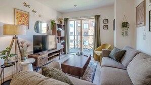 A modern living room with a flat-screen TV, sofa, coffee table, and a balcony with a view of buildings. - Host & Stay | Austin House (Norwich)