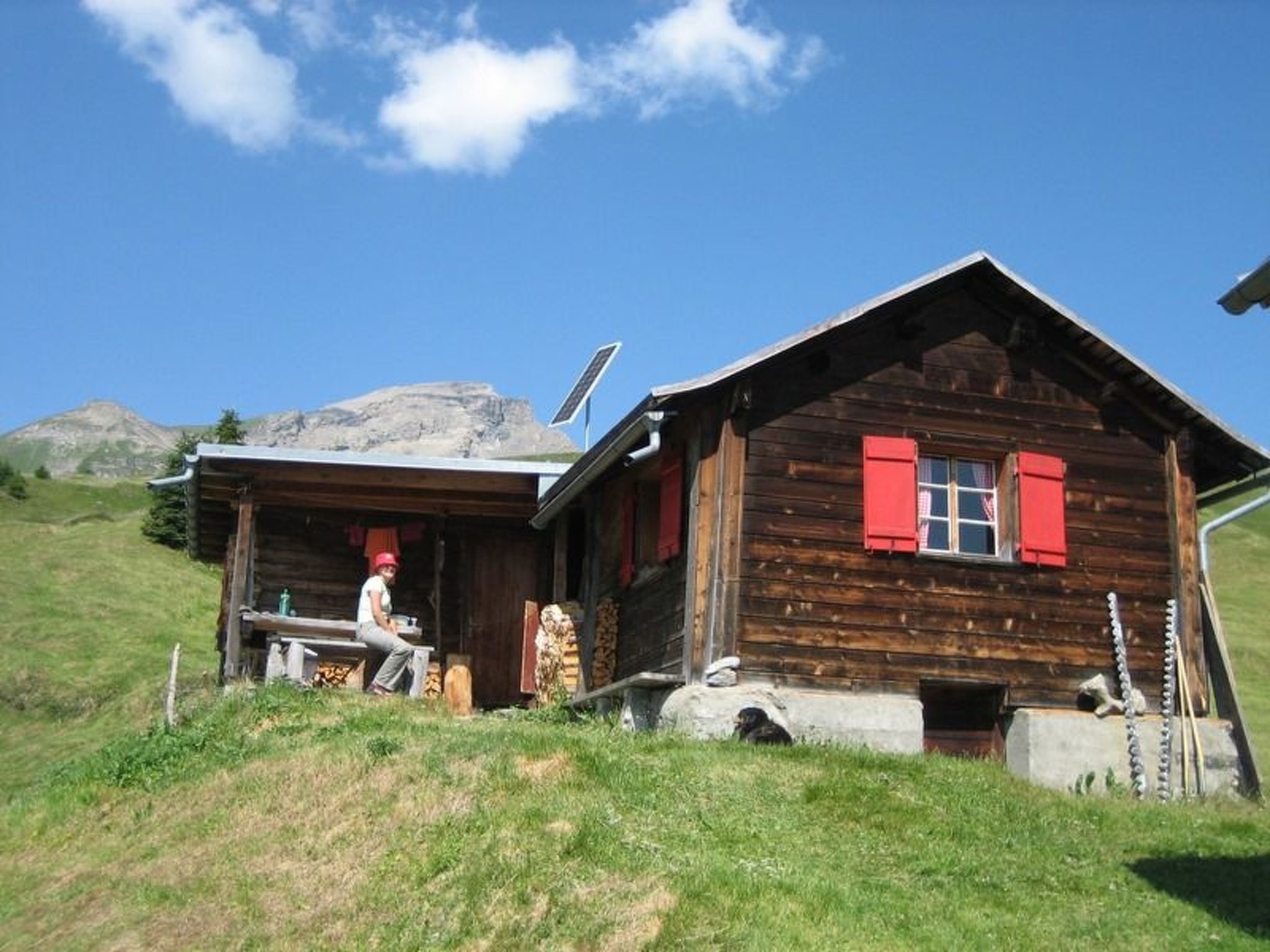 A wooden cabin with a sloped roof, a red door, and a person sitting outside.
