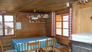 Une cabane en bois avec une table à manger, des chaises, un poêle et une fenêtre avec des rideaux.