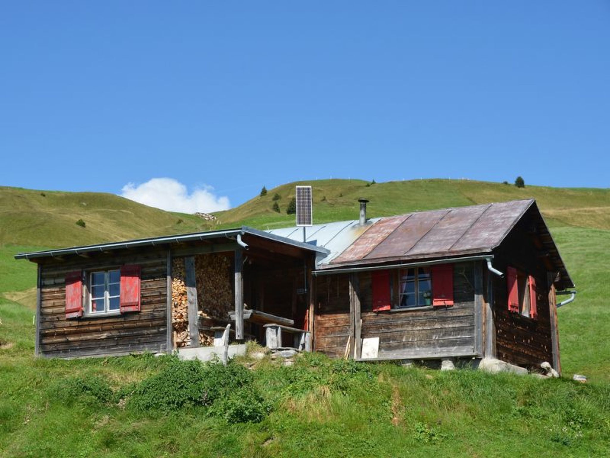 Une cabane rustique en bois, surmontée d’un toit en métal, située sur une colline herbeuse sous un ciel dégagé.