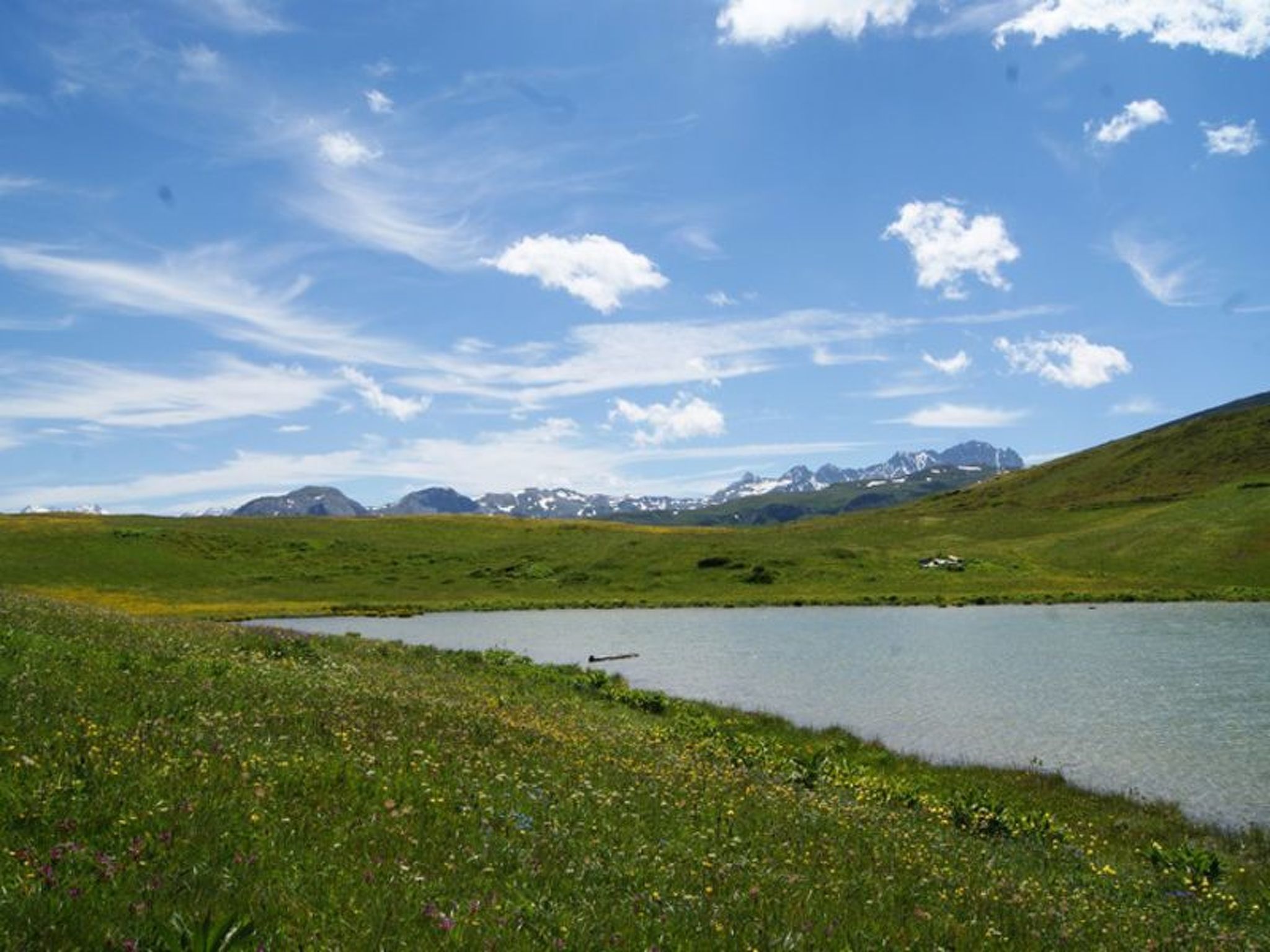 Un paysage naturel avec un lac, des prairies verdoyantes et des montagnes lointaines sous un ciel dégagé.