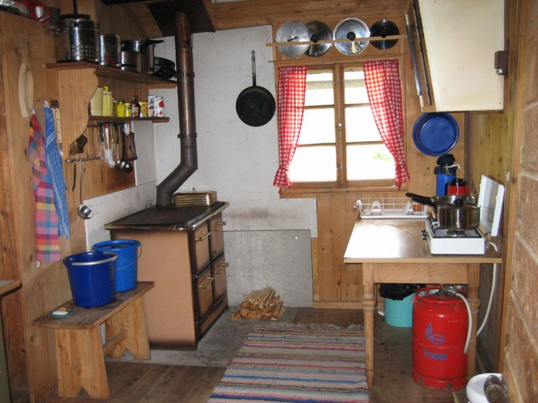 Una cuina senzilla amb un foc de llenya, una finestra amb cortines quadriculades i diversos utensilis i recipients de cuina.