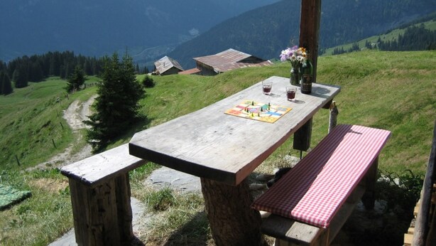 Une table de pique-nique en bois, recouverte d’une nappe à carreaux, avec une bouteille et des verres dessus, le tout placé devant un paysage montagneux.