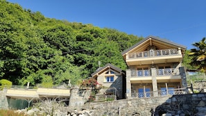 Ein zweistöckiges Steinhaus mit Balkonen, einer Steinmauer und einer bewaldeten Hügellandschaft im Hintergrund.