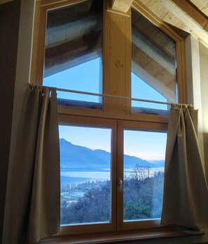 Ein Fenster mit Blick auf einen See und Berge, Vorhänge und Holzbalken.