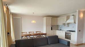 En moderne stue med en grå hjørnesofa, et spisebord med stole, et køkken med hvide skabe, en indbygget ovn og en pendel.